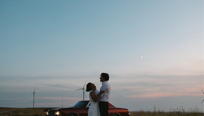 A man and woman hold each other in front of a vintage car during blue hour.