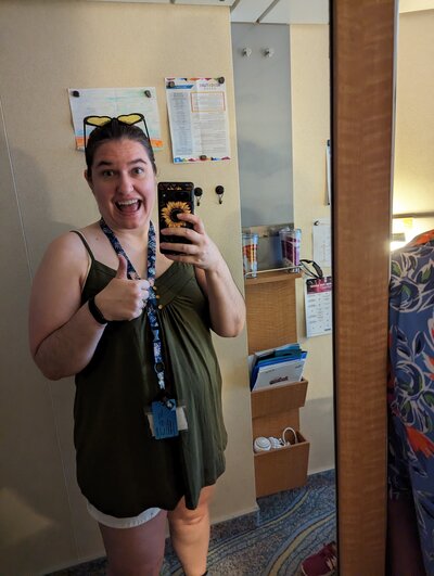 The travel advisor gives a thumbs up, wearing a cruise lanyard, yellow heart-shaped sunglasses, and a big goofy grin as she raises her cell phone.