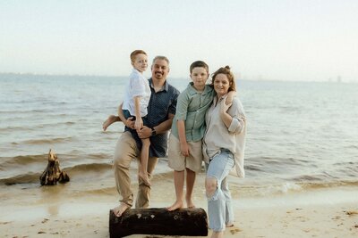 A family of four poses barefoot on a sandy beach near the water, with two adults and two children standing close together and smiling at the camera.