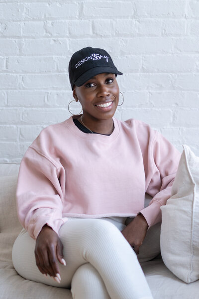 A casual branding portrait of a creative entrepreneur wearing a branded hat, seated in a light and airy studio against a white brick wall. Natural light highlights the relaxed and confident tone of the session.

