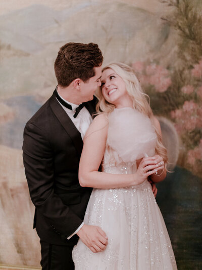 Kohnur Bride with soft glam makeup featuring rose gold eyeshadow, peach blush, and glossy nude lips, styled in romantic loose curls for an indoor ballroom wedding in California.