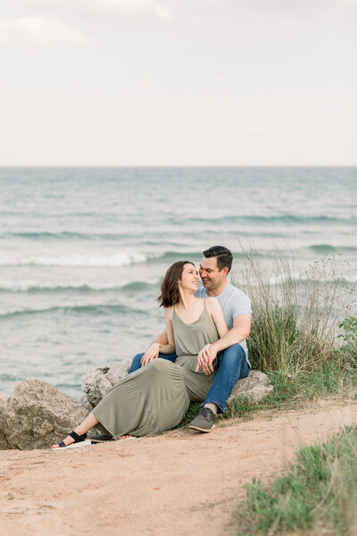 Couple sitting together on rocks with Lake Michigan in the background, beach grass nearby, laughing at each other