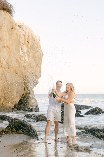 engaged couple pops champagne and sprays it on the beach during their engagement session in malibu captured by los angeles wedding photographer magnolia west photography