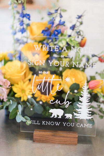 Acrylic guest book sign for a woodland-themed baby boy shower, displayed in front of a floral bouquet with yellow daisies, blue hyacinths, and greenery.