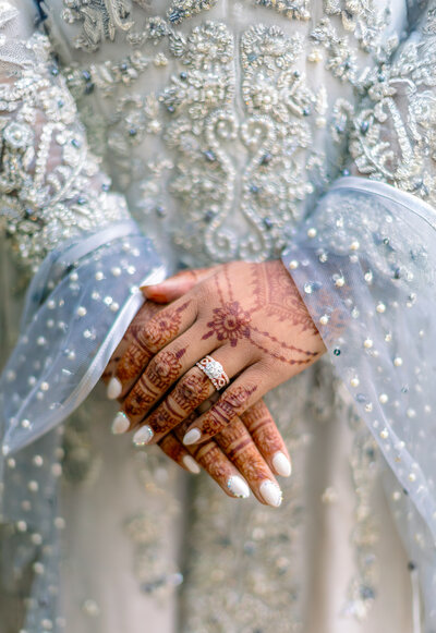 Close-up of a brides hands adorned with intricate henna designs and wearing an ornate ring. The attire features detailed silver embroidery and beadwork on sheer fabric, creating an elegant and decorative appearance.