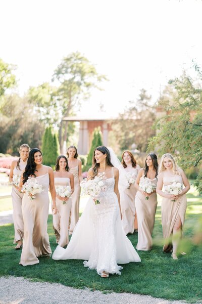 Bride smiling and walking with her bridesmaids in blush-toned dresses, each holding white bouquets, on a sunny garden path.