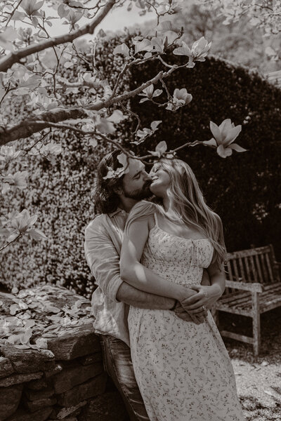 A man and woman stand under a tree blooming with flowers. They kiss.