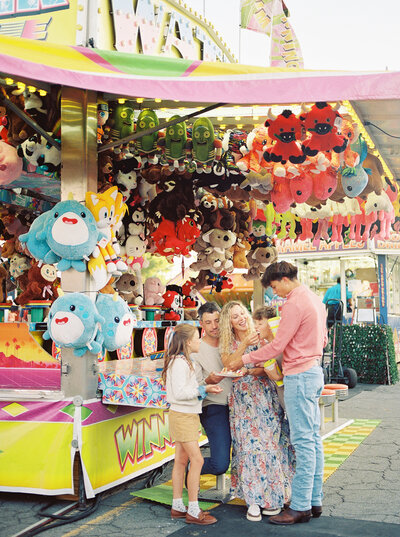 Family with tween and teenagers sit at a fair contentedly sharing a funnel cake under a canopy of prize toys captured by photographer Little Rock Bailey Feeler