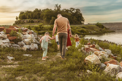 Dad in brown shirt and green pants walks barefoot holding his kids hands on walkway by a lake at sunset outdoors