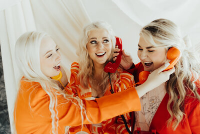 Kenzi in an orange suit, Sophie in a multicolored orange suit, and Marisa in a red suit holding phones to each others' ears