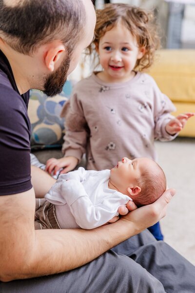 Dad holding newborn twin while older sister looks on. 