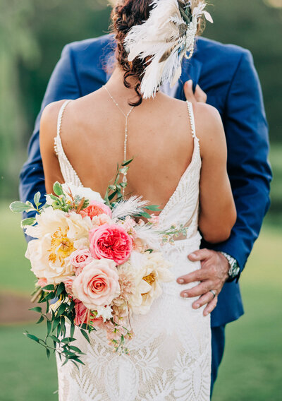 man in blue suit hugging woman in white dress facing away from the camera with pink and white bouquet