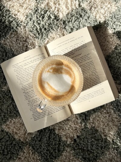 A glass coffee cup with frothy latte sits on an open book on a textured, patterned rug. Sunlight casts soft shadows, creating a cozy, relaxed mood.