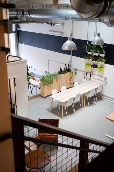 Modern industrial workspace with long communal table, white chairs, and greenery accents in open loft-style office.