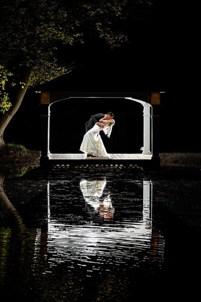 A groom dips his bride at night in front of a lake