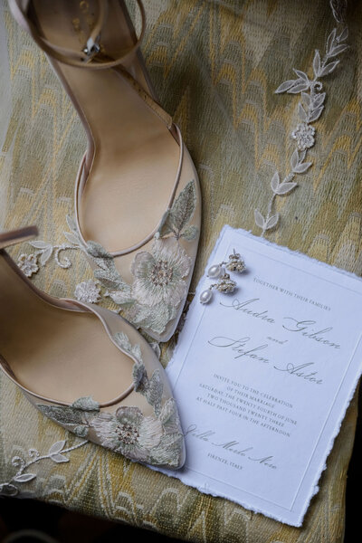 A pair of beige high-heeled shoes with intricate floral embroidery placed alongside a wedding invitation and pearl earrings on a textured green fabric background, exuding a romantic and sophisticated vibe.