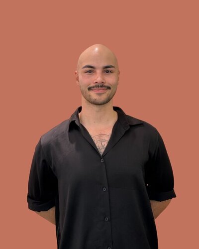 Bailey, Male hairdresser with styled short hair, dressed in a relaxed black outfit against a warm-toned backdrop.