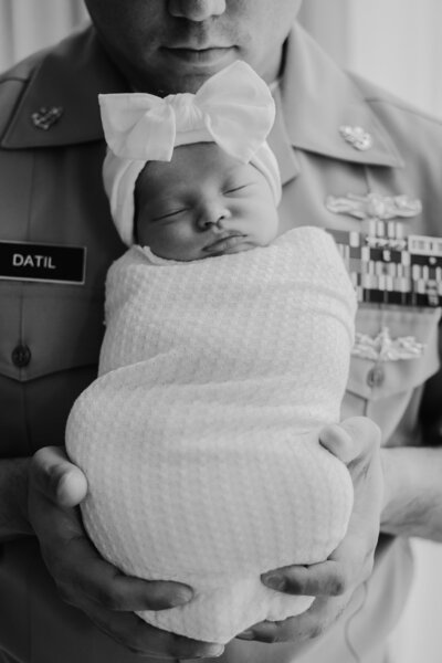 Black and white photo of dad in navy uniform holding baby girl wrapped in white blanket with white bow in moody lifestyle studio in Annapolis Maryland.