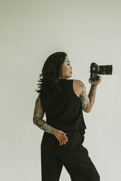 A tattooed woman dressed in black stands in profile, holding a camera in one hand and looking over her shoulder against a plain light background.