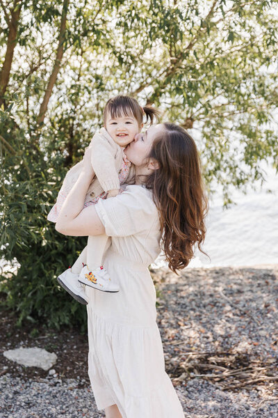Mom holding up toddler girl while kissing her on the cheek during their toronto family portrait session