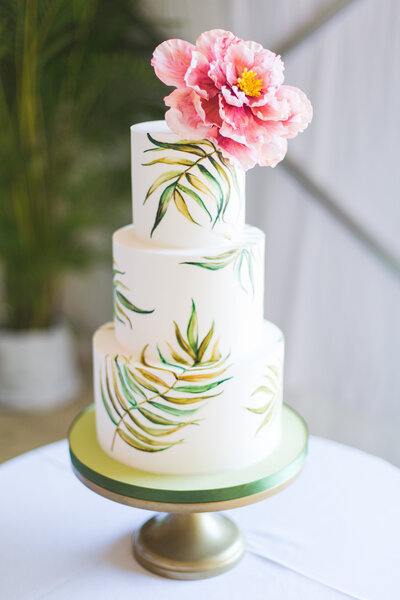Modern white tropical 3-tiered wedding cake with green palm branch design and large pink flower topper