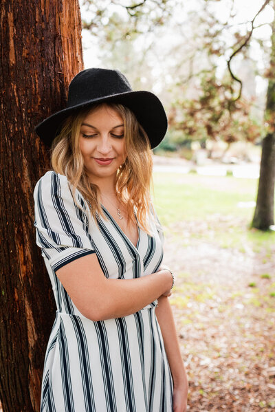 Girl wearing a white dress and black hat standing among evergreen tress in Point Defiance Park for senior photo session