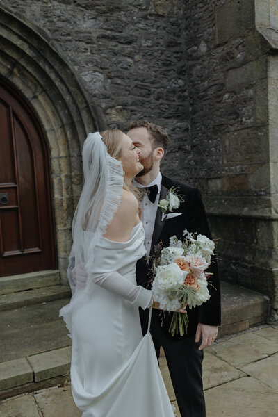 Bride and groom share a kiss outside historic church doorway, bouquet in hand, with timeless Luis Calow editorial (clitheroe) tones.