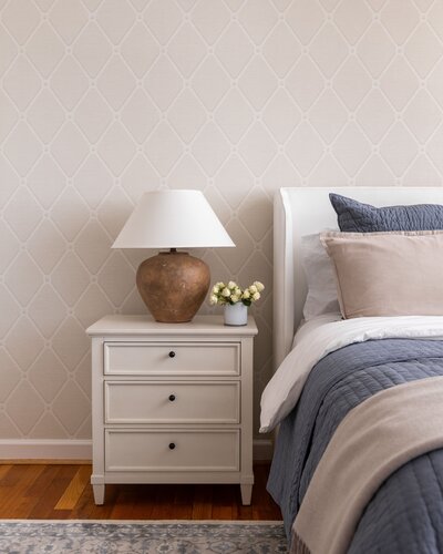 Bedroom with a mustard yellow upholstered headboard, white bedding, and a small gold and glass side table. The wall features a scenic mural depicting trees and classical architecture in muted tones.