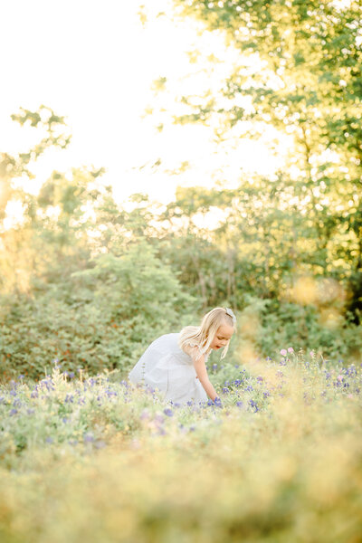 The picture captures a serene and enchanting moment in a field of vibrant bluebonnets. A young girl, dressed in a light blue spring dress, stands amidst the sea of delicate blue flowers, her face aglow with a radiant smile. The setting sun casts a warm, golden hue over the scene, creating a soft and dreamy atmosphere.  The girl's delicate features are accentuated by the natural beauty that surrounds her. Her hands are gently plucking the bluebonnets, as if she is carefully selecting the most beautiful ones to admire. The forest backdrop complements the girl's elegant attire.  The photographer, Bri Sullivan, has masterfully captured the essence of this spring photo session, using a light and airy style that enhances the natural beauty of the scene. The image is a testament to the beauty of nature and the simple joys of childhood, a moment frozen in time that evokes a sense of wonder and tranquility.  The overall composition of the photograph is both visually stunning and emotionally captivating. The girl's pose, the vibrant colors of the bluebonnets, and the warm glow of the sunset all come together to create a truly breathtaking image that will be cherished for years to come.