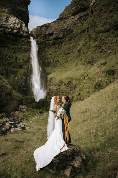Couple exchanges vows at the foot of a waterfall, Iceland’s lush greenery surrounding them.