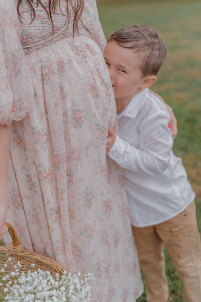 A young boy gently hugs his mother's pregnant belly as he smiles at the camera.