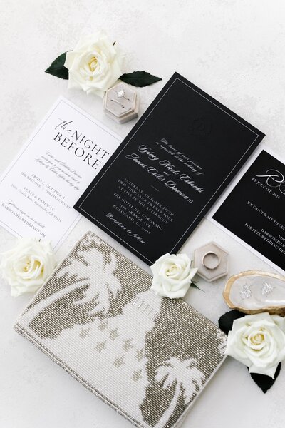 Flat lay of wedding invitation suite with roses, ring box, clutch, and jewelry on a light surface. This wedding was planned and designed by Thater Made, a luxury wedding and event producer based in San Diego, CA.