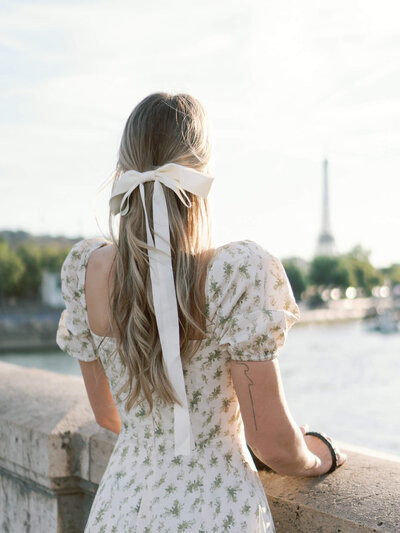 Woman with long blonde hair tied with a ribbon, wearing a floral dress, looking toward the Eiffel Tower in Paris.
