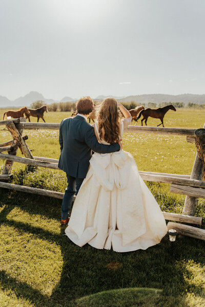 bride and groom standing in a field next to a wooden fence watching horse run