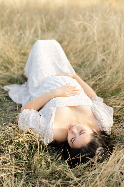 A pregnant mother lays in the grass in a beautiful dress while she holds her pregnant stomach.