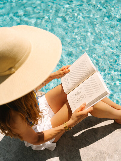 Woman in a straw hat sitting and reading a book beside a pool
