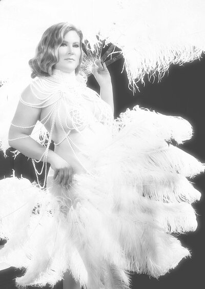 A woman is draped in pearls with white feather fans and photographed like a burlesque performer like Christina Aguilera.
