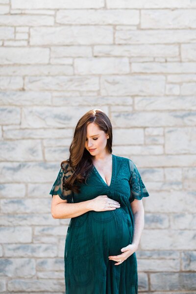 Pregnant woman in a green dress holding her belly while standing in front of a stone wall.