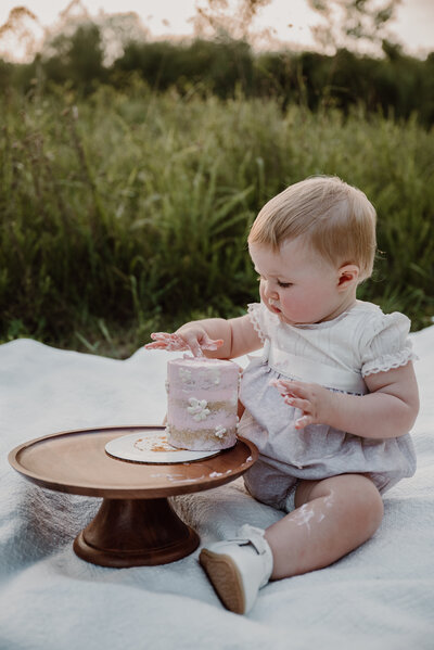 One year old baby girl wearing white and pink heirloom bubble sitting in a sitting on a white blanket with a brown cake stand smashing a pink cake.