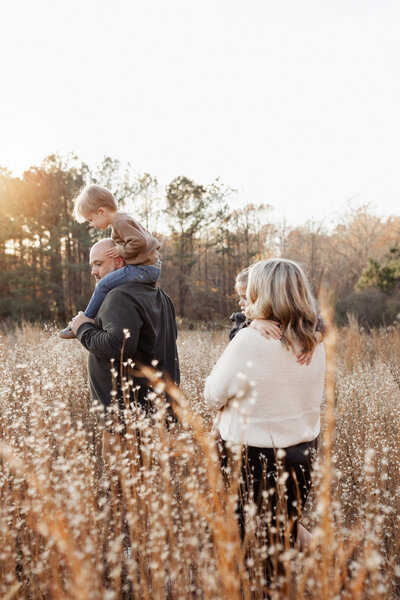 A family walks through tall brown grass that is glowing with the sunset at Hyde Farm Park. 