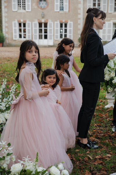 Four young flower girls in blush pink dresses stand in a row during an outdoor wedding ceremony, beside a woman in a black suit.