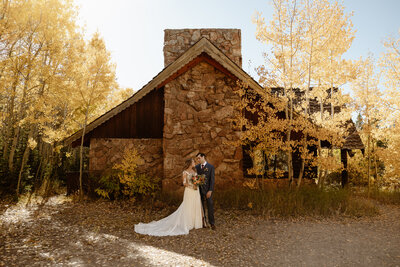 image of bride and groom standing in front of a cabin in their wedding attire surrounded by golden aspen trees taken by colorado elopement photographer