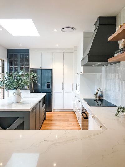 This modern kitchen in Newcastle features bespoke interior design with marble countertops, a black range hood, and sleek white cabinets. A skylight illuminates the space, highlighting the elegance of luxury home design enhanced by beautiful wooden flooring.