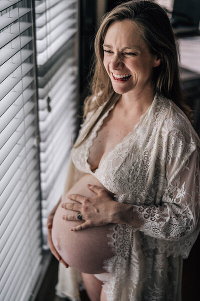 A pregnant mama holds her belly and laughs by her living room window while wear a gorgeous lace gown during an in home lifestyle pregnancy photo session.