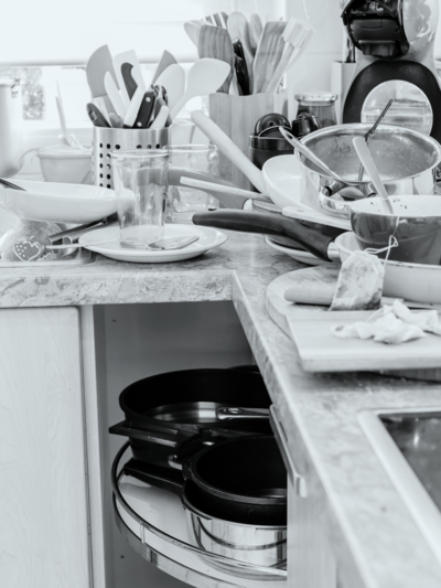 Black and white photo of a cluttered kitchen counter filled with dirty dishes, pans, and utensils.