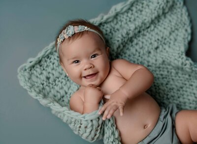 Baby girl in teal diaper cover, laying on back, grabbing blanket and smiling for her 6 month milestone photo. Milestone baby photo taken at Atlanta Maternity photo studio in Woodstock GA.