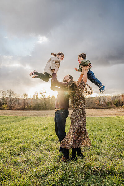 mom and dad stand back to back and hold kids in air at sunset