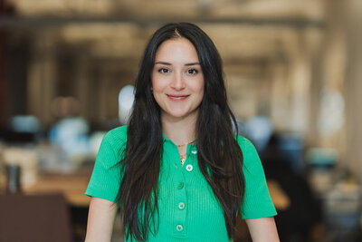Professional headshot of young woman in green blouse with long dark hair, smiling in office setting