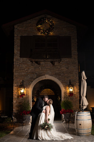 Nighttime portrait of couple kissing, the bride holding her bouquet down by her side, in front of the entrance to the main building at Childress Vineyards in Lexington, NC. Christmas bows are adorned on lanterns and a lit wreath add warmth to the image.