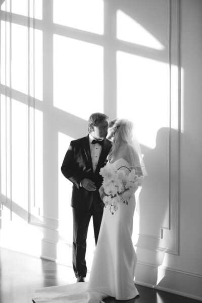 Bride and groom in formal attire standing in soft window light with geometric shadows, capturing an elegant wedding moment indoors.